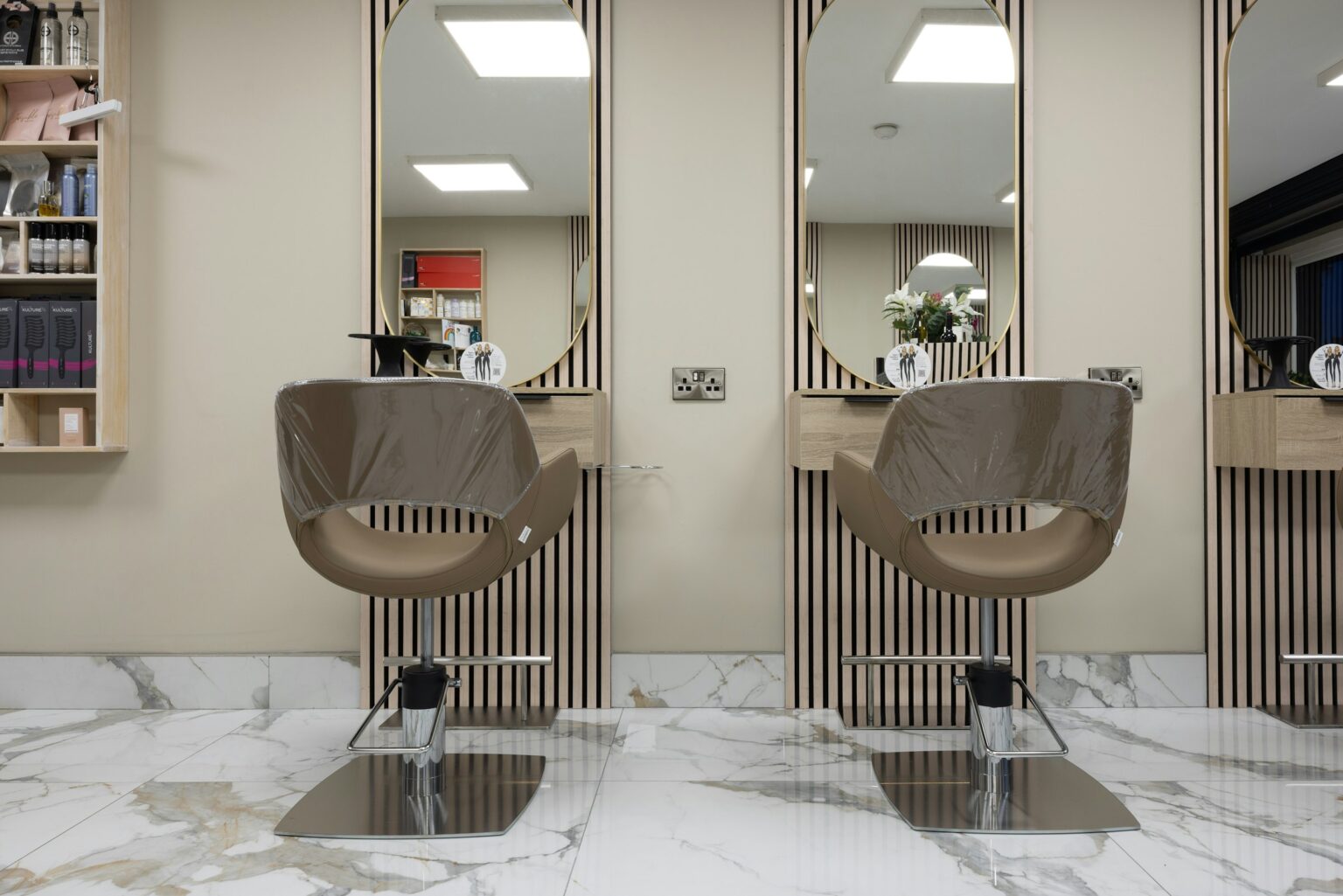 Two styling chairs facing tall oval mirrors in a softly lit salon with a marble floor